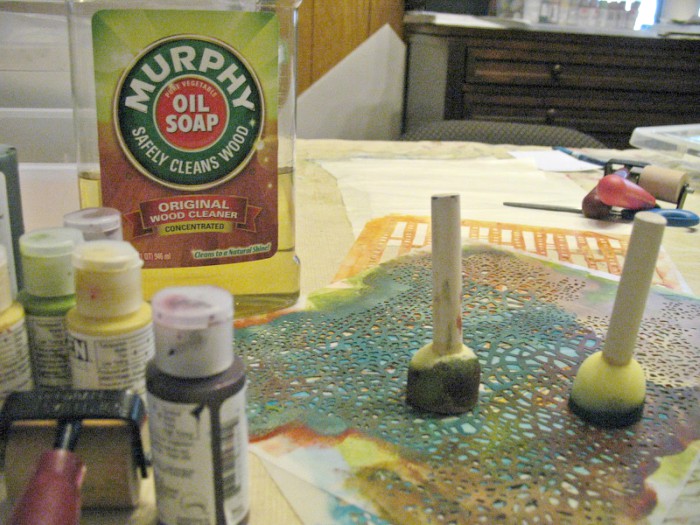 A bottle of Murphy's Oils Soap, bottles of paint, sponge brushes and stencils on a table.