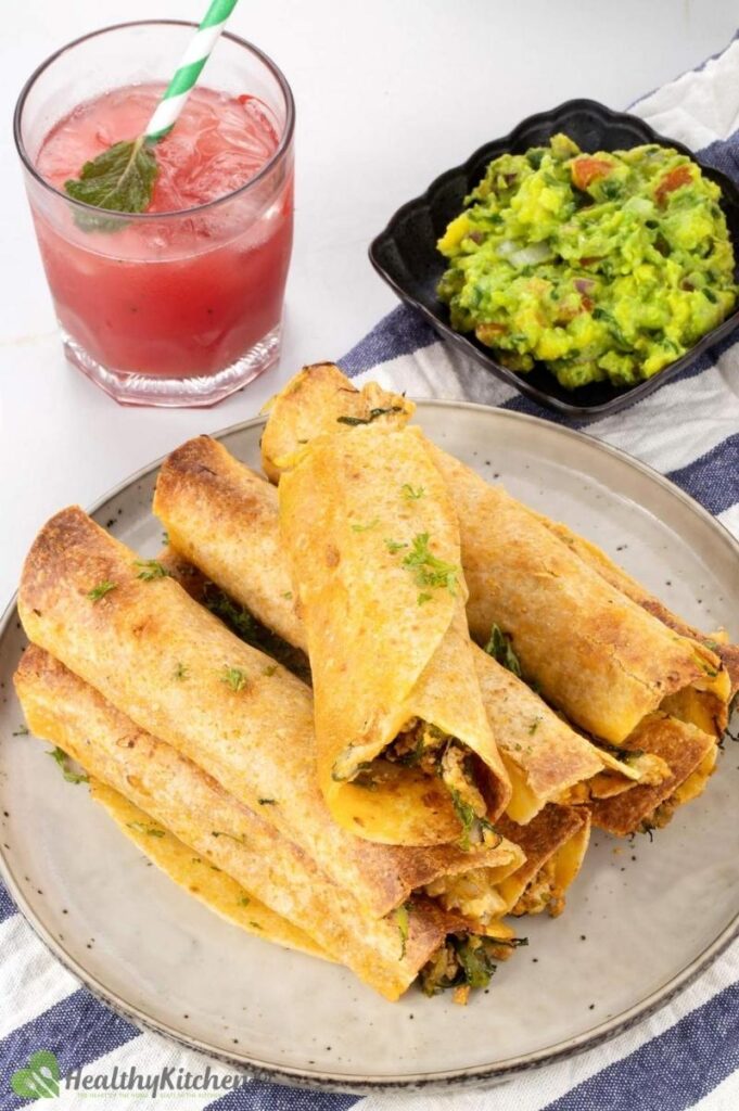 Baked chicken taquitos on a plate with a bowl of guac and a cocktail