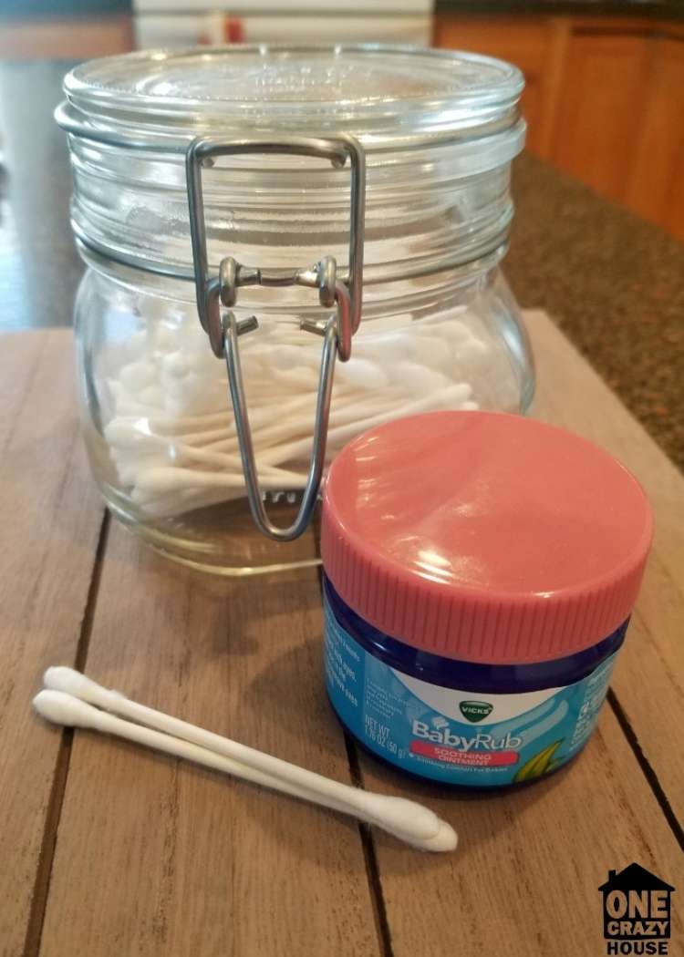 Must Try Vicks VapoRub hacks! In foreground are 2 cotton swabs. On their right is a small jar of Vicks BabyRub, and behind it is a large glass jar containing cotton swabs. All items are placed on a wooden table. 