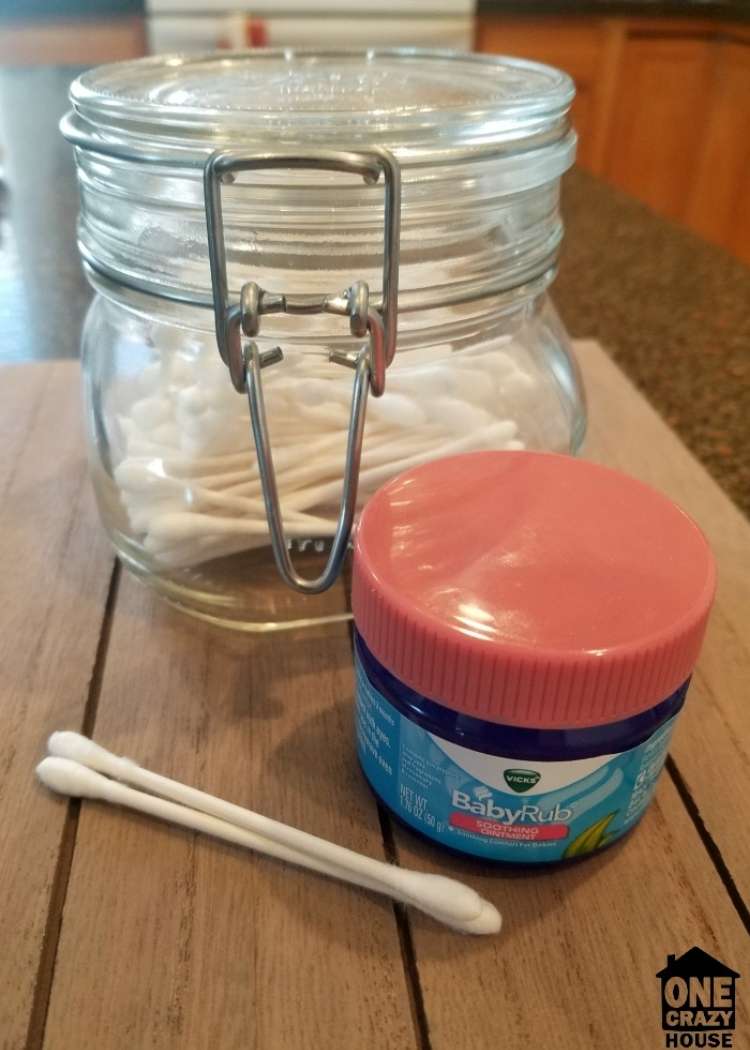 Must Try Vicks VapoRub hacks - 2 cotton swabs in the foreground. On their right is a small jar of Vicks BabyRub, and behind it is a large glass jar containing cotton swabs. All items are placed on a wooden table. 