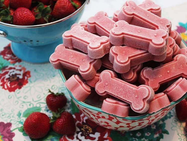 Bowl of strawberry and yogurt frozen treats in shape of dog bones on table. Bowl of strawberries in background with strawberries scattered around bowl.