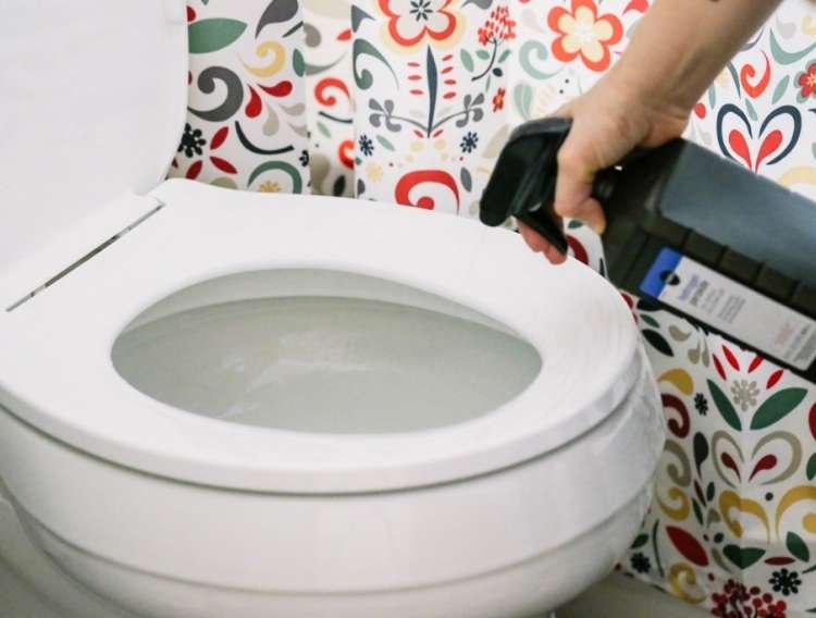 hand holding Toilet bowl being sprayed with hydrogen peroxide with flower shower curtain in background