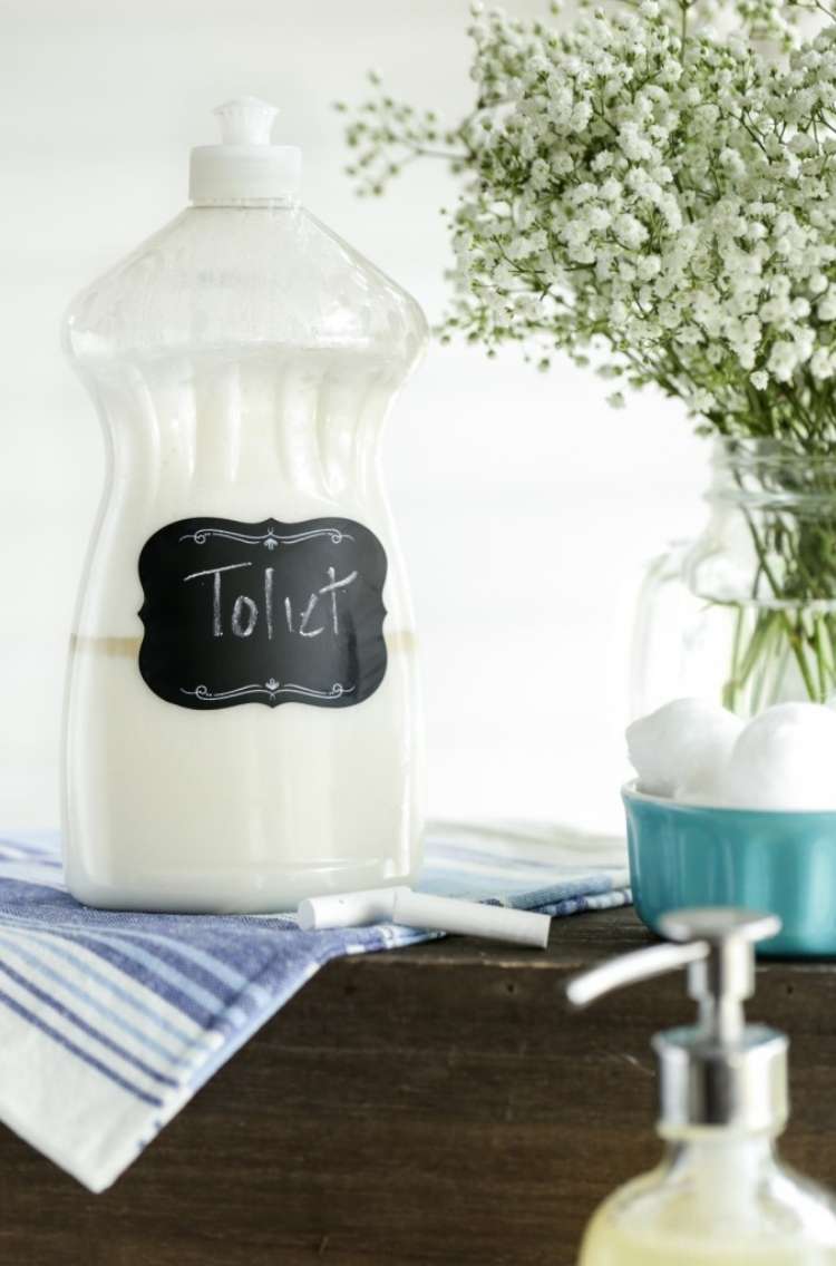 Dishsoap bottle labeled toilet sitting on a towel next to a bowl of homemade toilet cleaning bombs with flowers in the background in a glass vase and a soap dispenser pump in the foreground