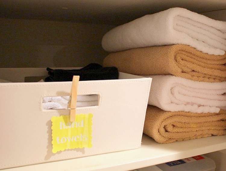 Onecrazyhouse Linen Closet Organization Shelf in a linen closet with a plastic container with a clothespin attached to the top holding a label. Stack of towels next to it