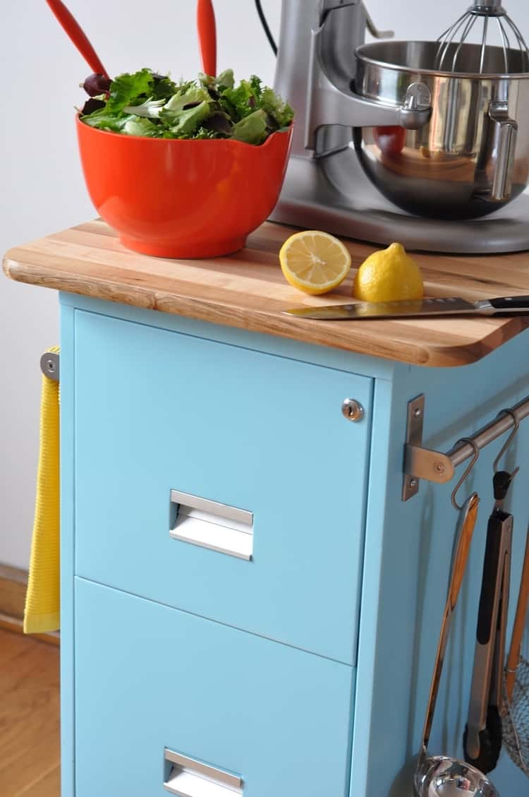 An orange bowl containing a vegetable salad next to 2 pieces of a sliced lemon, a kitchen knife, and mixer on a make over light blue kitchen cart 