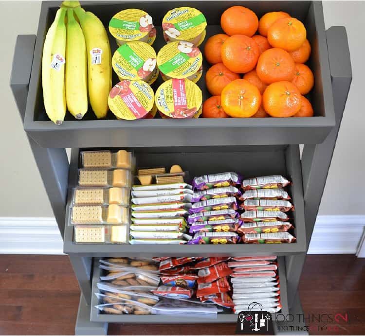 14 - Snack Organization Station on 3 tears with top one with bananas, yoghurt, clementines, 2nd tier different snacks, bottom cookies and snacks