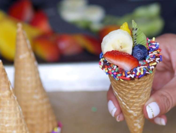 OneCrazyHouse Party Foods hand holding a waffle cone filled with fruits with sprinkles on the outside of the cone