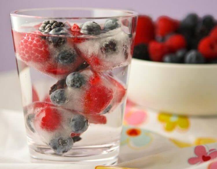 Ice cubes with frozen strawberries and blueberries inside of a clear glass with a bowl of berries in the background