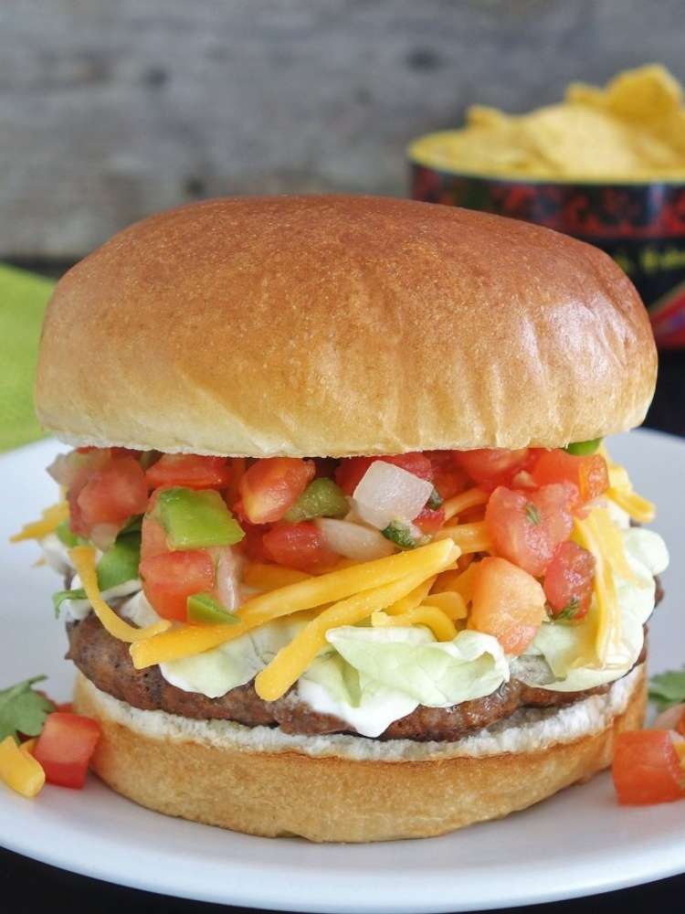 A taco burger topped with taco sauce, tomatoes, jalapenos, cheddar, lettuce, and onions.