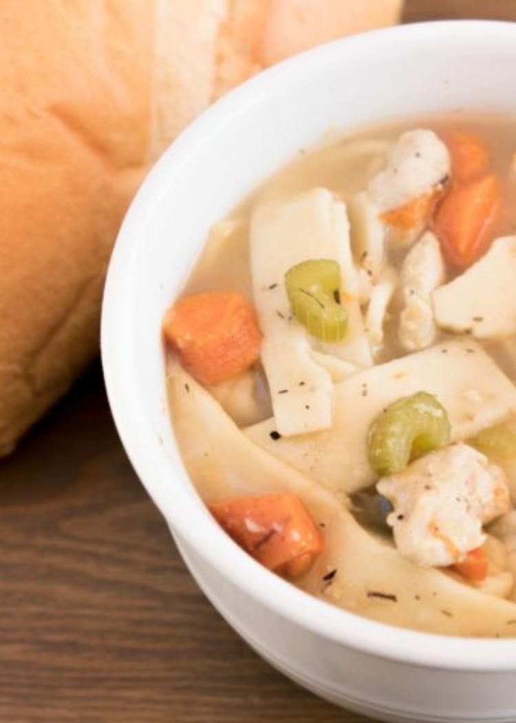 Chicken Noodle Soup - soup in white bowl with carrots, celery and flat short noodles. Bread love in background.