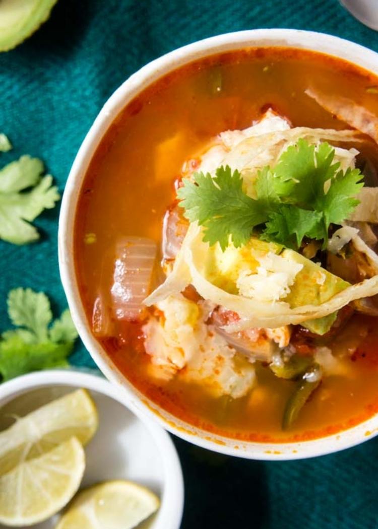 Red Tortilla Soup with lots of vegetables in soup. Garnished with lush parsley. With green tablecloth and small bowl of sliced lime in the background.