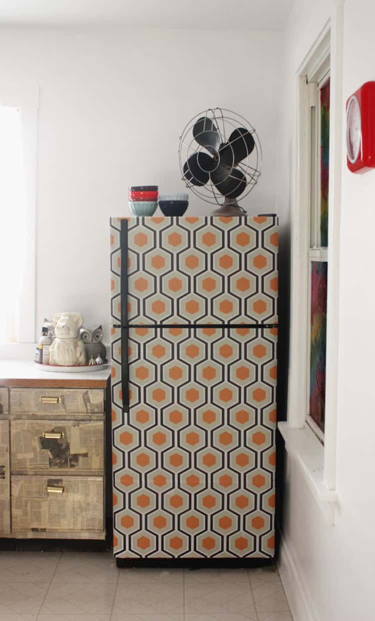 cabinet on left, and a fridge on the right with a fan on top and bowls, the fridge is wallpapered with a gold, white, and tan print color