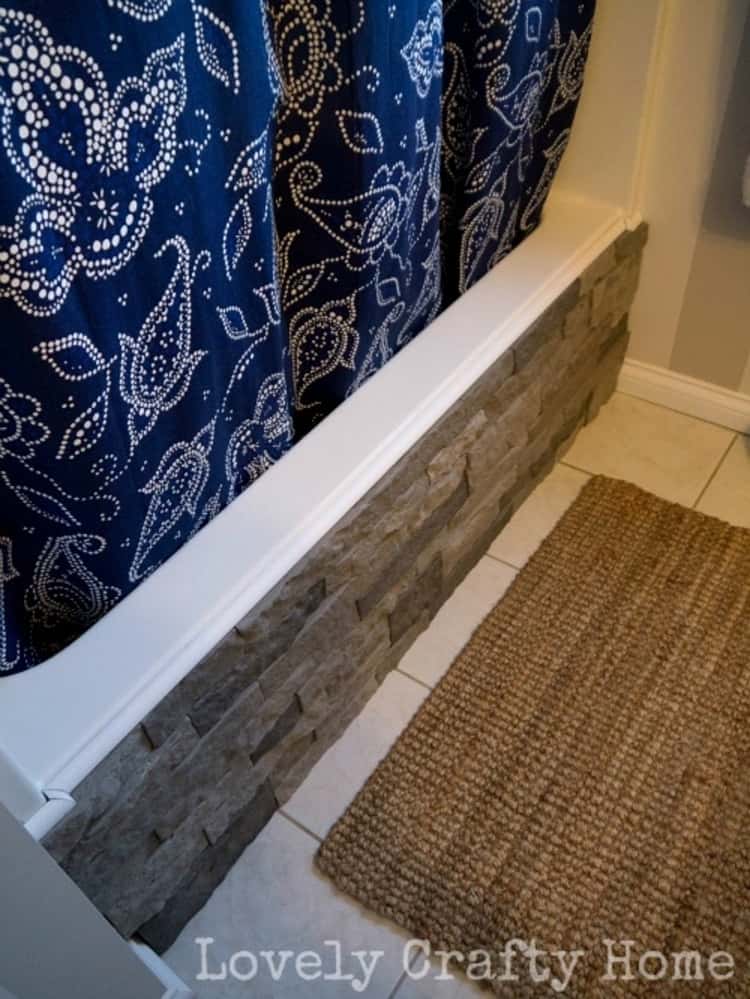 front of bathtub covered with grey airstone, blue print curtain to cover shower and a rug in front.