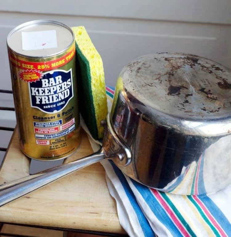 dirty stainless steel pot turned upside down with bar keepers friend sitting next to it and a green and yellow sponge in between.