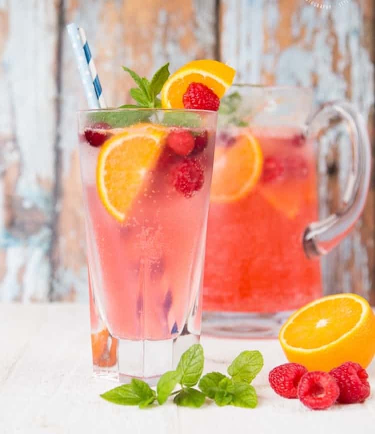 A glass and pitcher of garnished cranberry, raspberry, and orange sparkling cooler mocktail