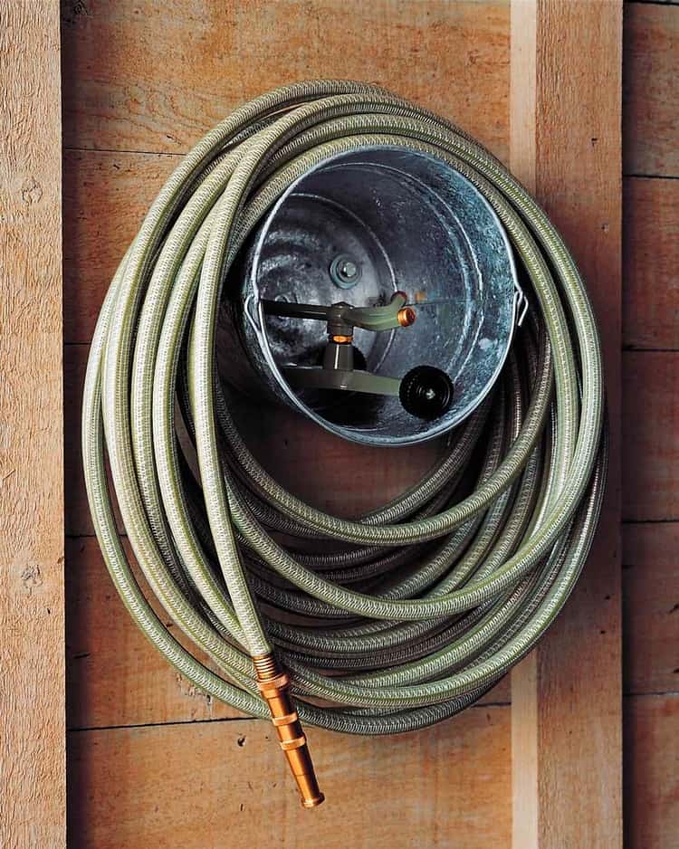 garden hose wrapped around a metal bucket hanging on the wall with hose attachment placed inside of bucket.