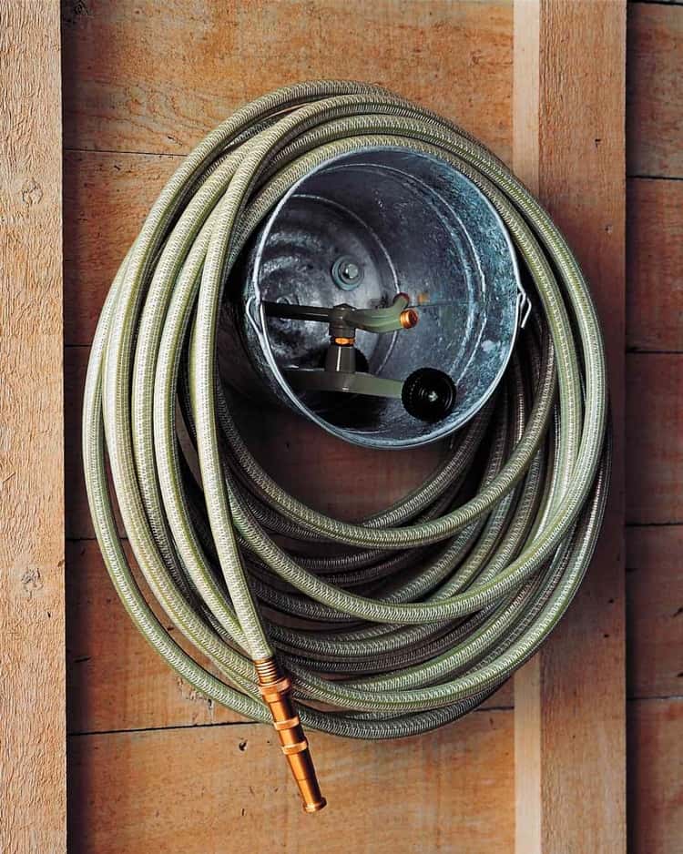 garden hose wrapped around a metal bucket hanging on the wall with hose attachment placed inside of bucket.