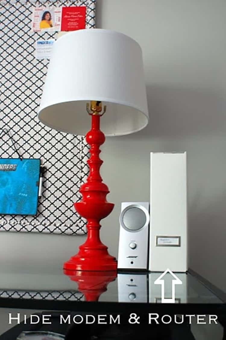 red lamp with white shade and next to it there's a magazine cover hiding a router/modem sitting on an end table