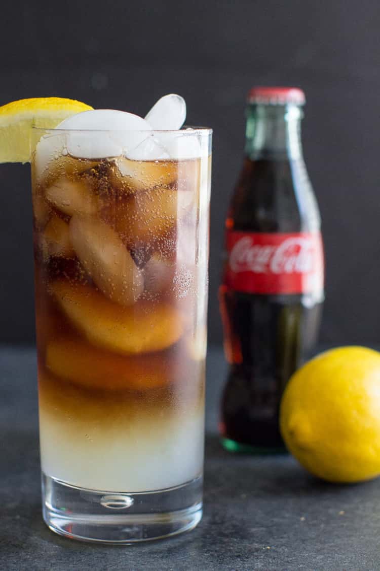 a glass of the Lemon Spezi mocktail, a bottle of Coca Cola, and a lemon. 