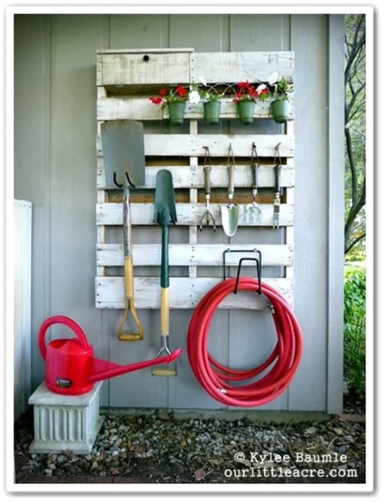 a wooden pallet hung on a wall and modified to hang your garden tool including flower pots, shovels, hand rake, hose and a plastic water can sitting next to it