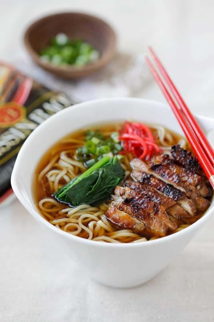 A bowl of ramen noodles in soy sauce with pieces of chicken topped with lemongrass and two chopsticks