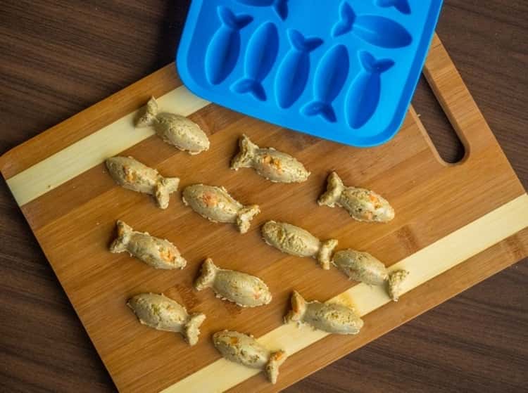 fish-shaped homemade salmon and sweet potato soft cat treats on a wooden cutting board next to a silicone fish mold