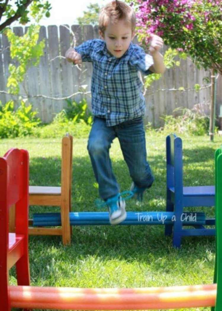 uses for pool noodles- a child jumping over pool noodles in a backyard obstacle course made out of chairs with pool noodles through the legs