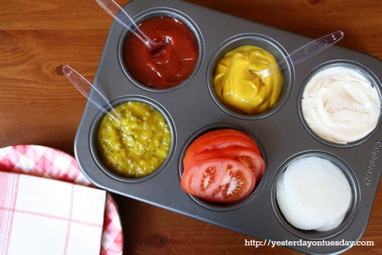 A muffin tray filled with a variety of condiments and vegetables with spoons for everyone to help themselves 