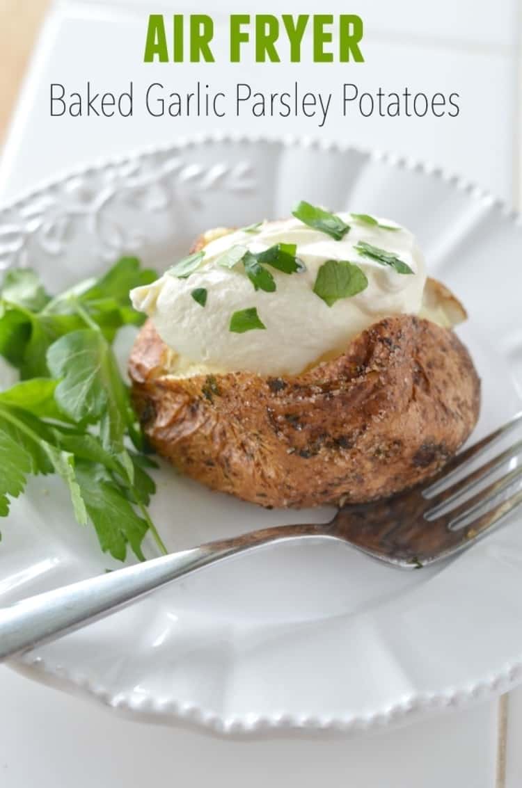 Air fryer baked potato topped with sour cream and chives on a white plate with a fork and a sprig of parsley on the side.