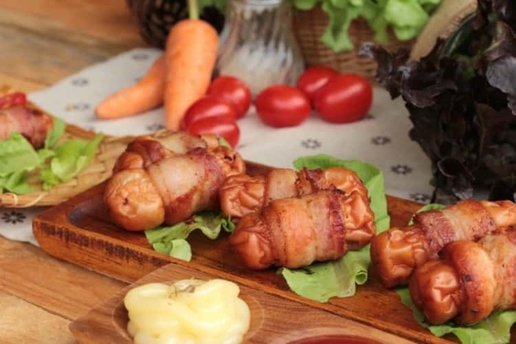 Bacon-wrapped air fryer- sausages on wooden tray with cherry tomatoes, carrots and pat of butter in a small wooden dish on the side.