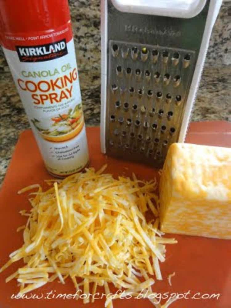 Grated cheese, cheese block, cheese grater and a can of cooking spray placed on a board.