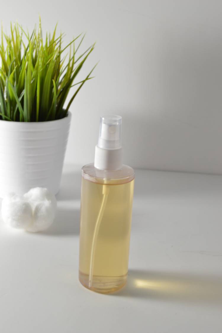 An image of homemade diy skin toner in a spray bottle. Background is a white counter. Next to the toner is a cotton ball and a green plant in a white pot.