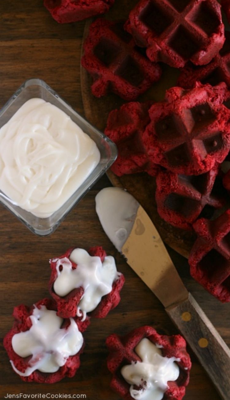 a pile of red velvet waffle cookies on a cutting board with a side of cream frosting in a bowl with a spreading tool