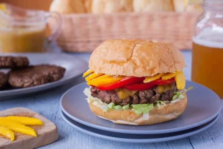 A beef burger topped with lettuce, jerk suace, grilled red peppers, and grilled mango slices.